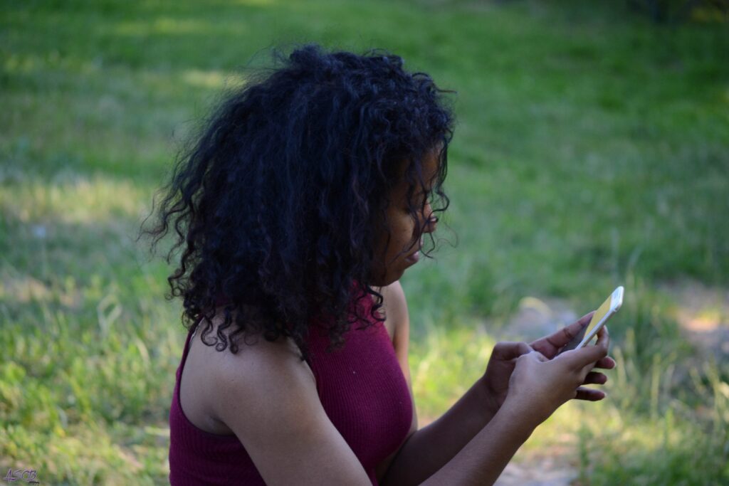 Side view of woman with natural dark curls sitting outside in a park, looking down at her phone.