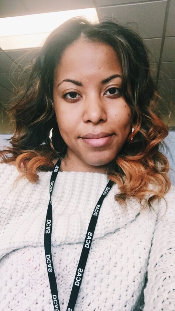 Woman with loosely curled ombre hair wearing a white knit sweater and work lanyard, showing signs of heat styling on color-treated hair.