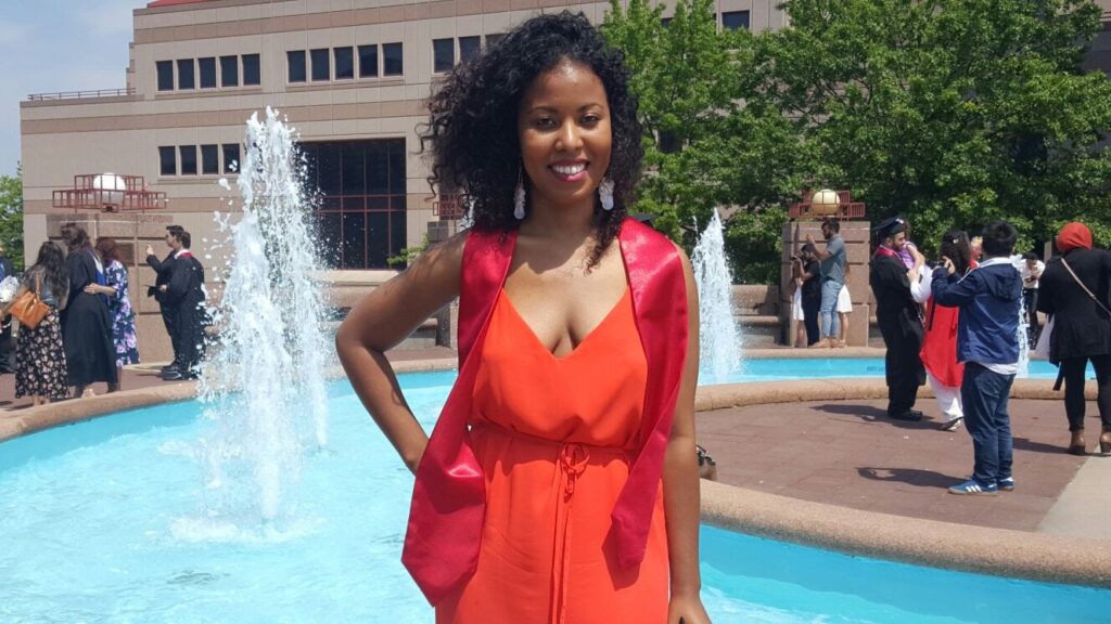 Woman wearing a red graduation sash and orange jumpsuit smiling in front of a fountain after college graduation.