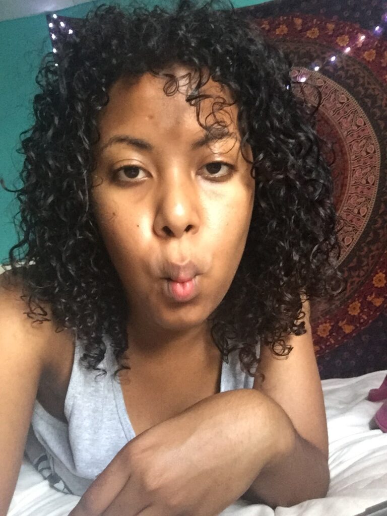 Woman with defined, slightly wet curls making a fish face in a casual, relaxed moment at home.