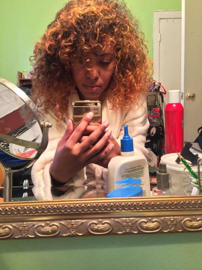 Mirror selfie of a woman with honey-blonde curly hair in a white robe, showcasing her natural hair care routine with visible products on the vanity.