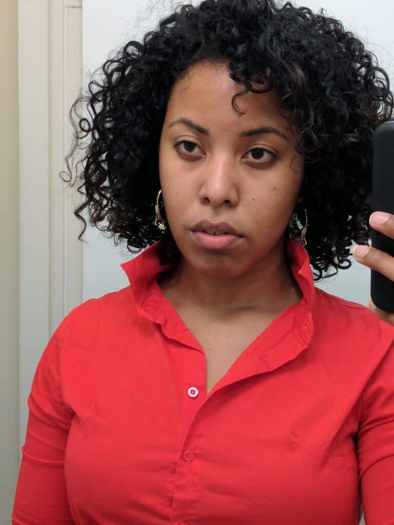 Woman with short, natural curly hair looking directly at the camera in a red collared shirt, showcasing early stages of hair regrowth after damage