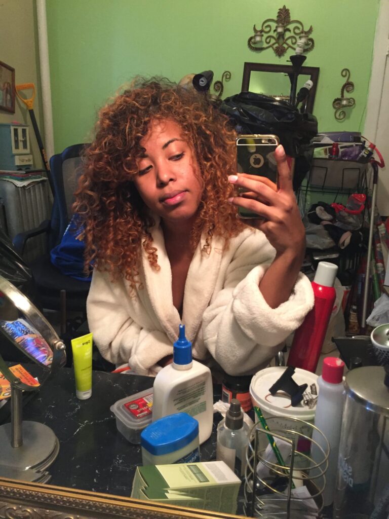 Natural curly-haired woman taking a relaxed mirror selfie in a cozy at-home setup, highlighting her textured hair and daily styling products.