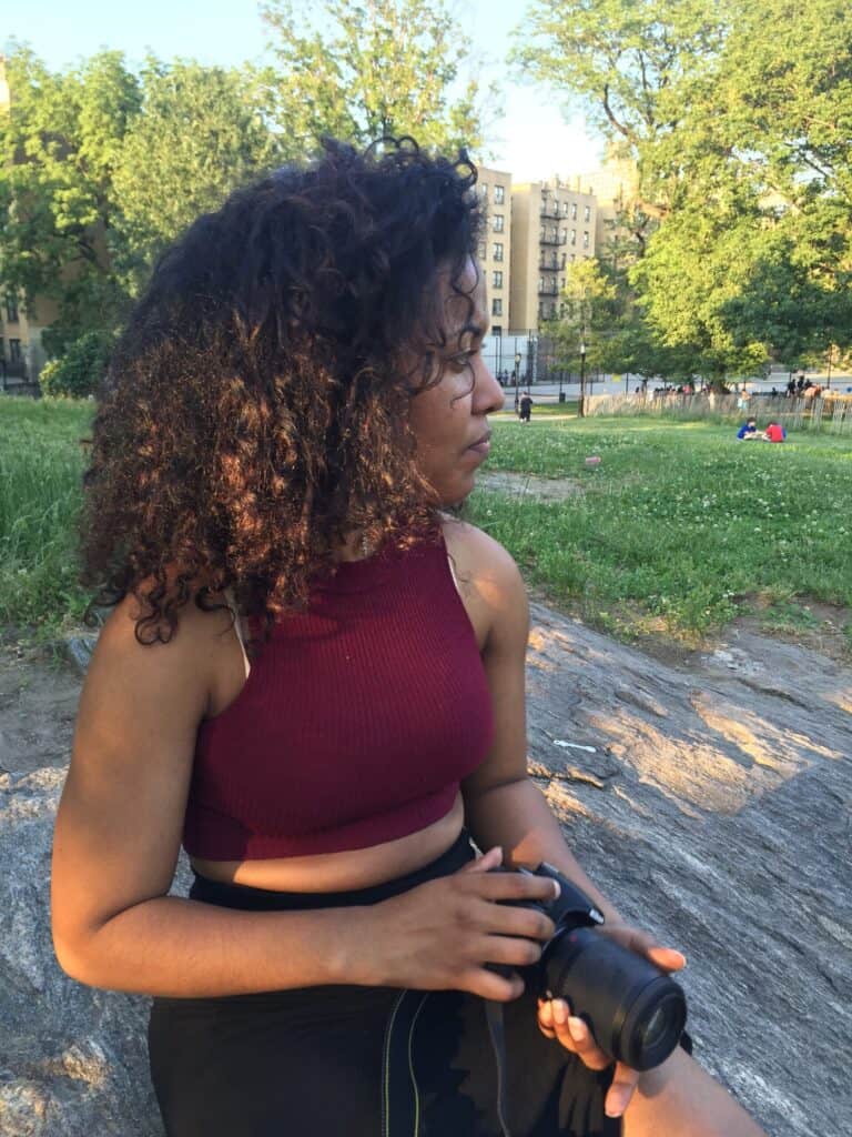 Woman with curly hair sitting in a park wearing a maroon tank top, looking away.