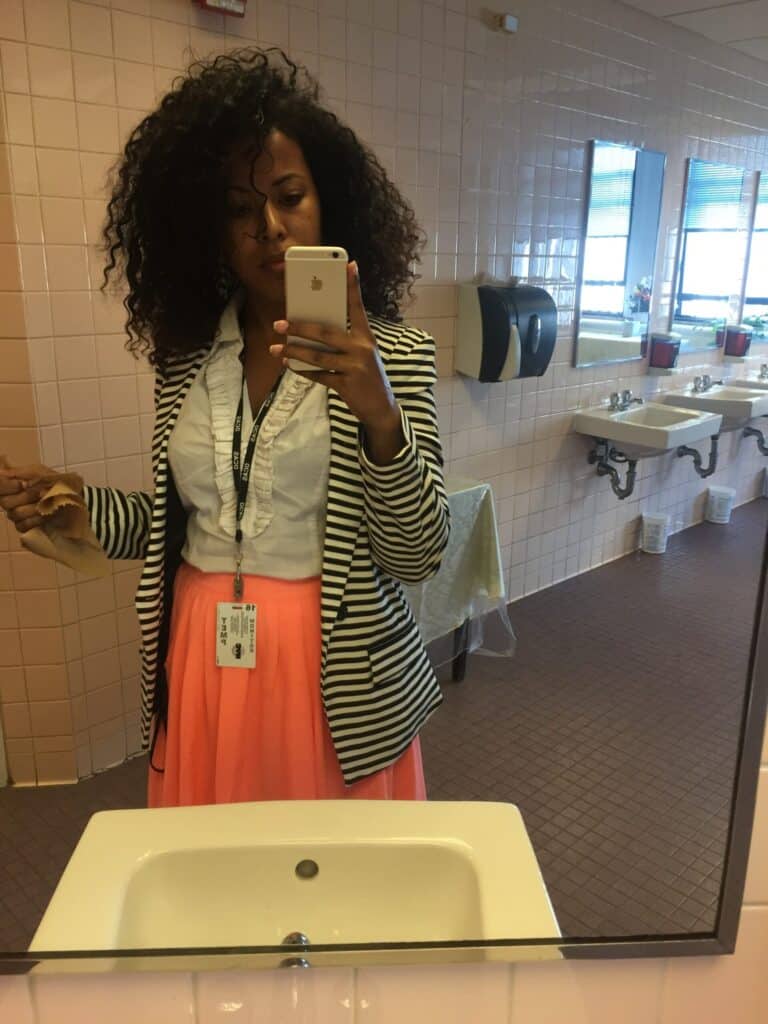 Woman with curly hair wearing a pink dress and striped blazer, taking a mirror selfie in a public restroom.