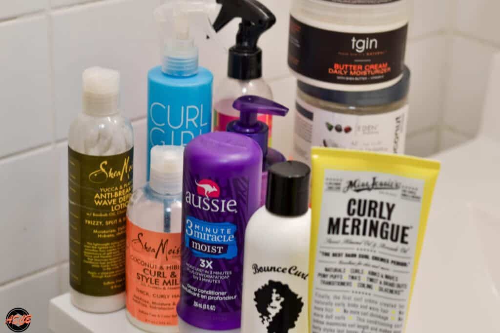 Collection of various curly hair care products including brands like Shea Moisture, Aussie, Bounce Curl, Miss Jessie's, and TGIN arranged on a bathroom ledge.