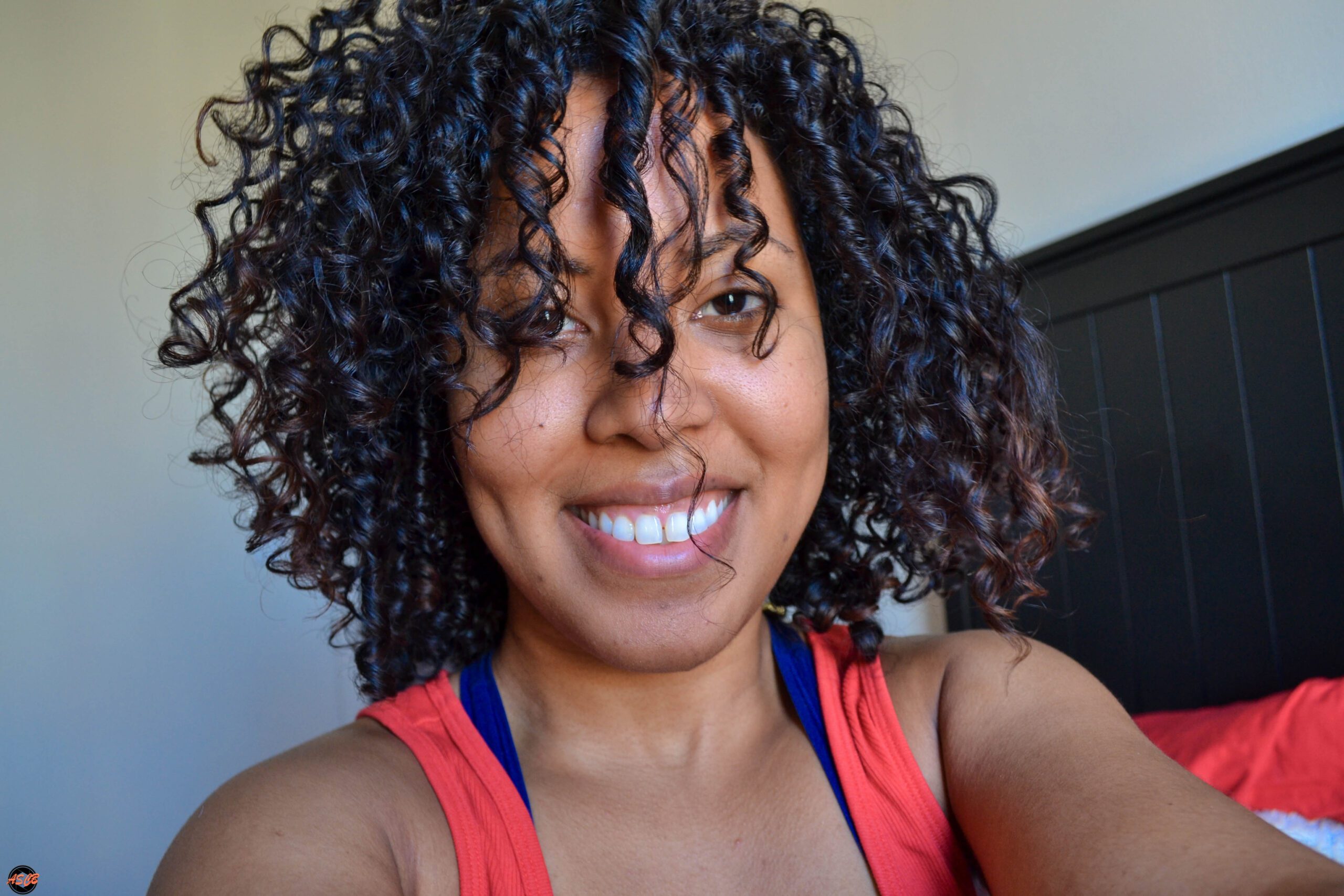 Close-up of a woman with defined, moisturized curls and minimal makeup, smiling in bright indoor light.