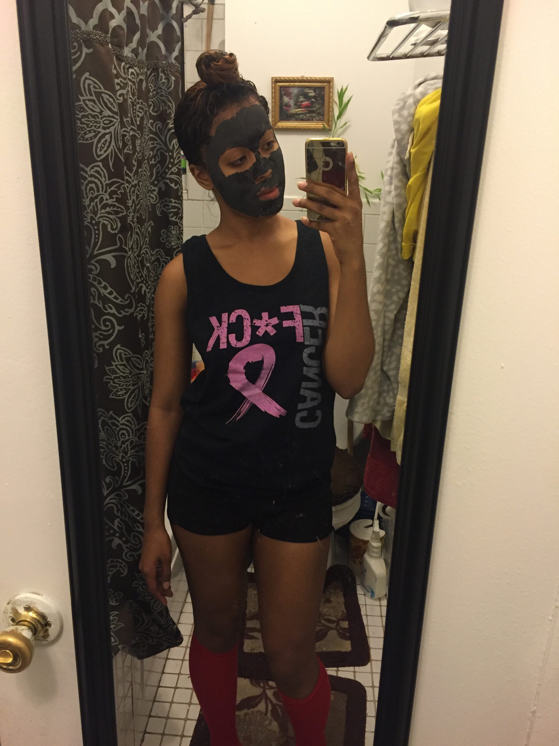 Woman taking a mirror selfie while wearing a black bentonite clay face mask, mid-treatment in a casual at-home setting.