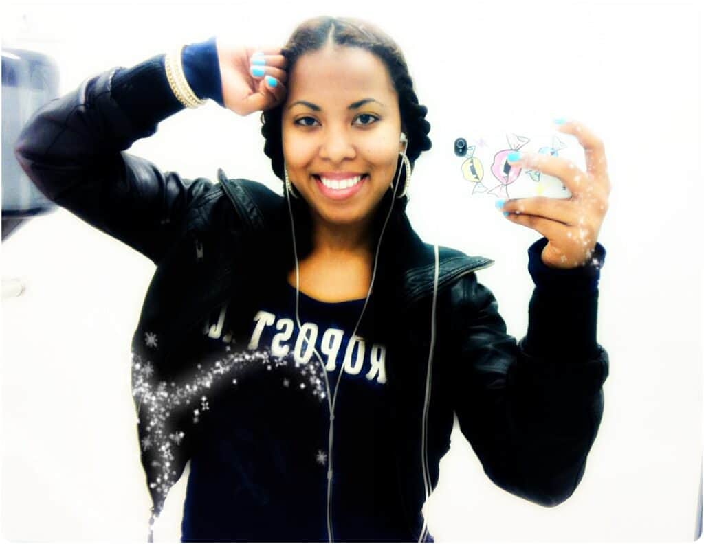 Smiling woman wearing two-strand braids as a protective hairstyle, holding her phone for a selfie. She’s dressed in a leather jacket and hoop earrings, with bright blue nail polish.