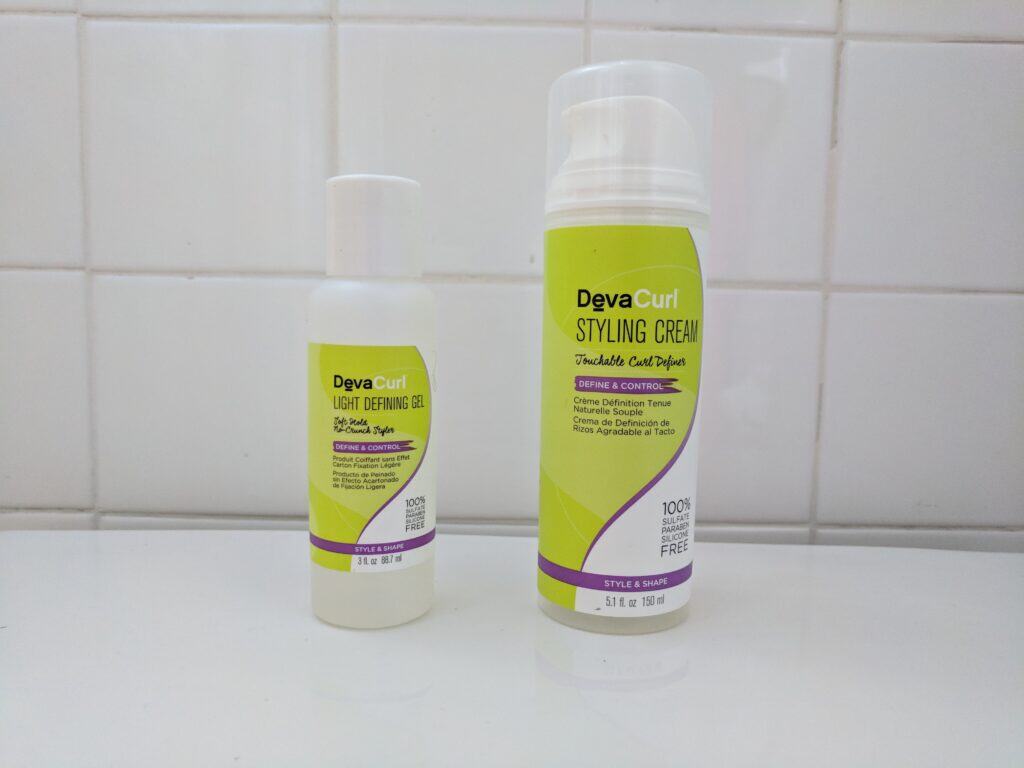 DevaCurl Styling Cream and Light Defining Gel bottles standing side by side on a white tile bathroom counter.
