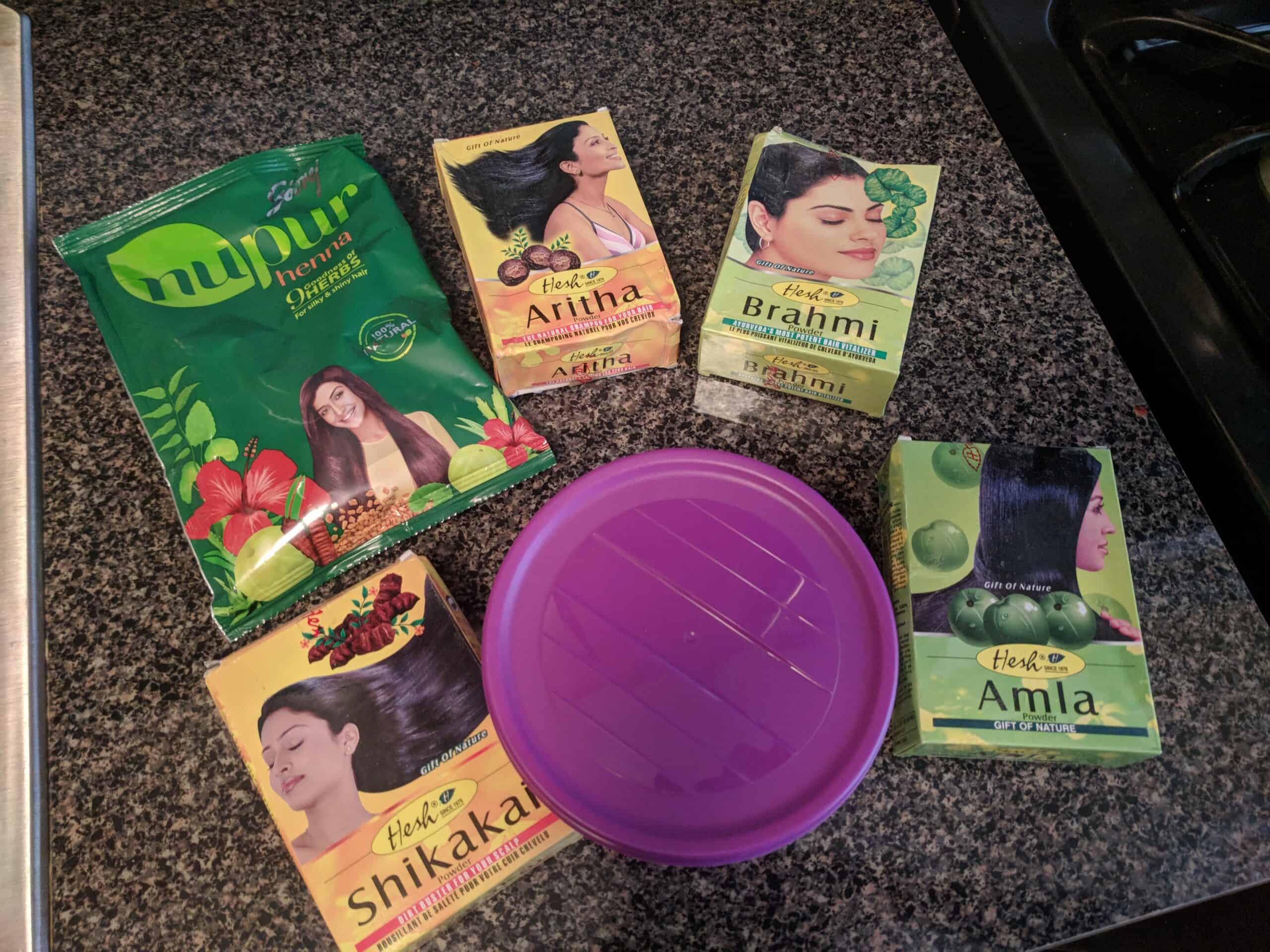 A collection of Ayurvedic hair powders displayed on a kitchen counter, including Nupur Henna, Hesh Aritha, Brahmi, Shikakai, and Amla, alongside a purple mixing bowl for preparing DIY hair masks.