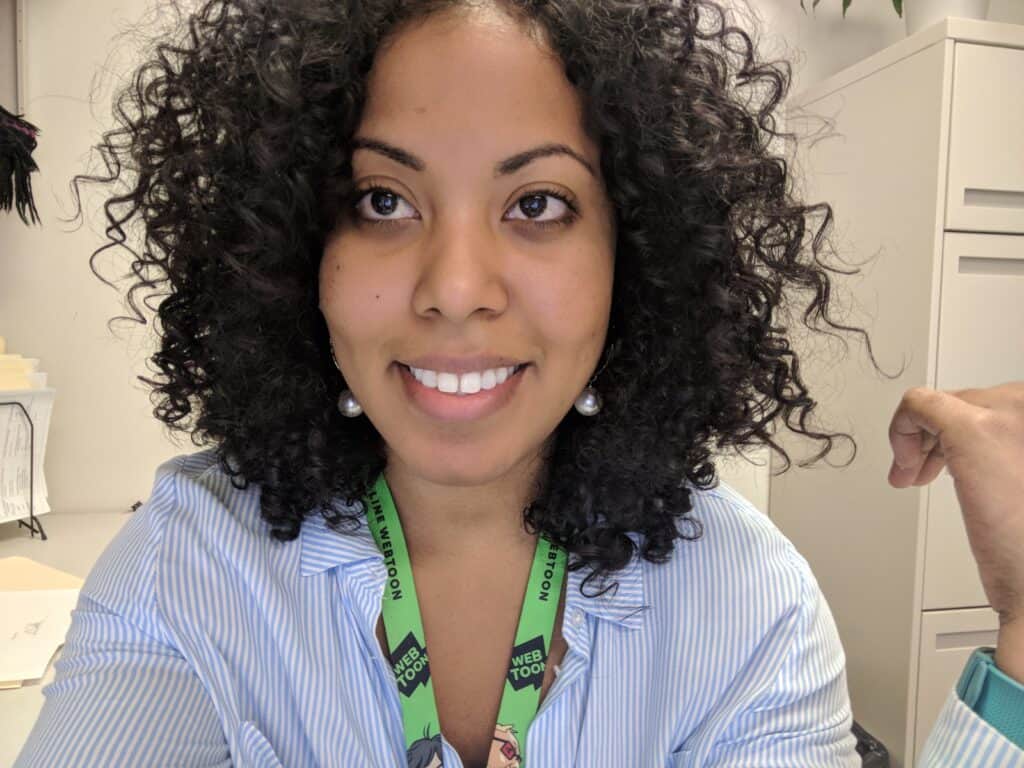 Close-up of a woman with dark, defined curls and a radiant smile, wearing a striped shirt and green lanyard.