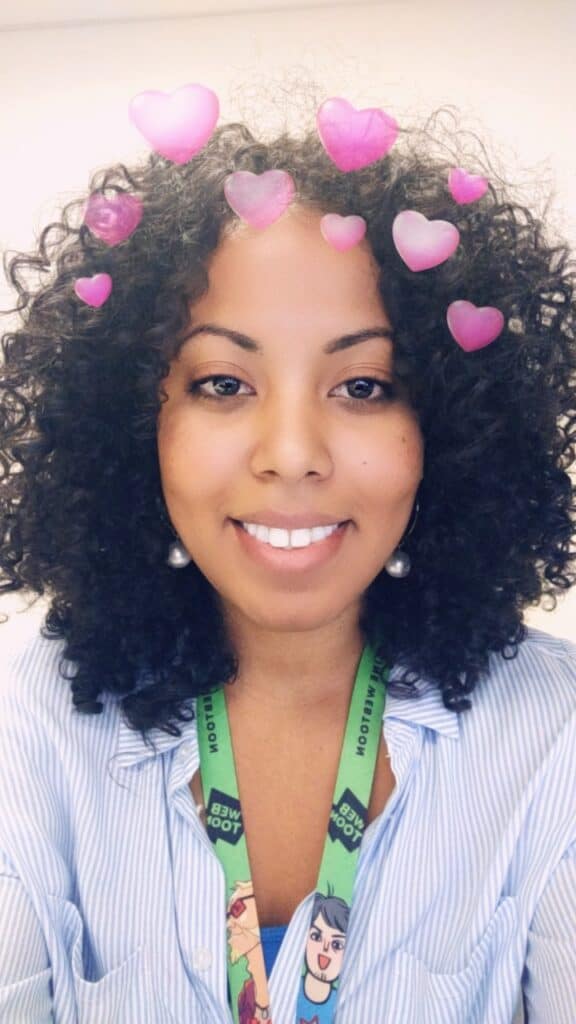 Smiling woman with soft, voluminous curls, wearing a striped shirt and green lanyard, with pink heart filter over the image.