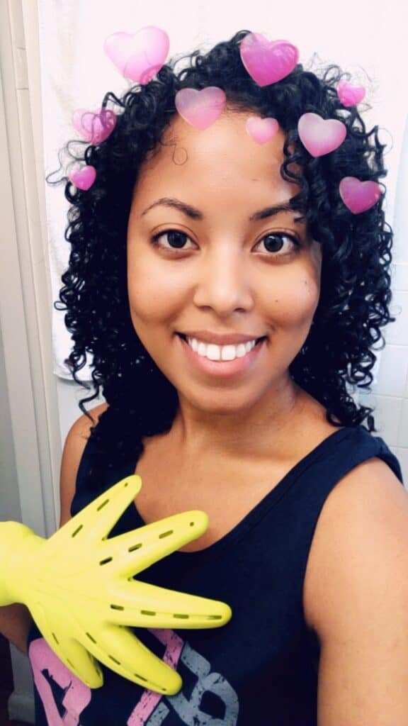 Smiling woman with freshly defined curls, holding a yellow hair tool, with pink heart filter over her head.