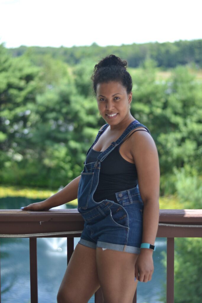 Smiling woman in denim overalls posing on a balcony with a scenic green landscape in the background, wearing a fitness tracker.