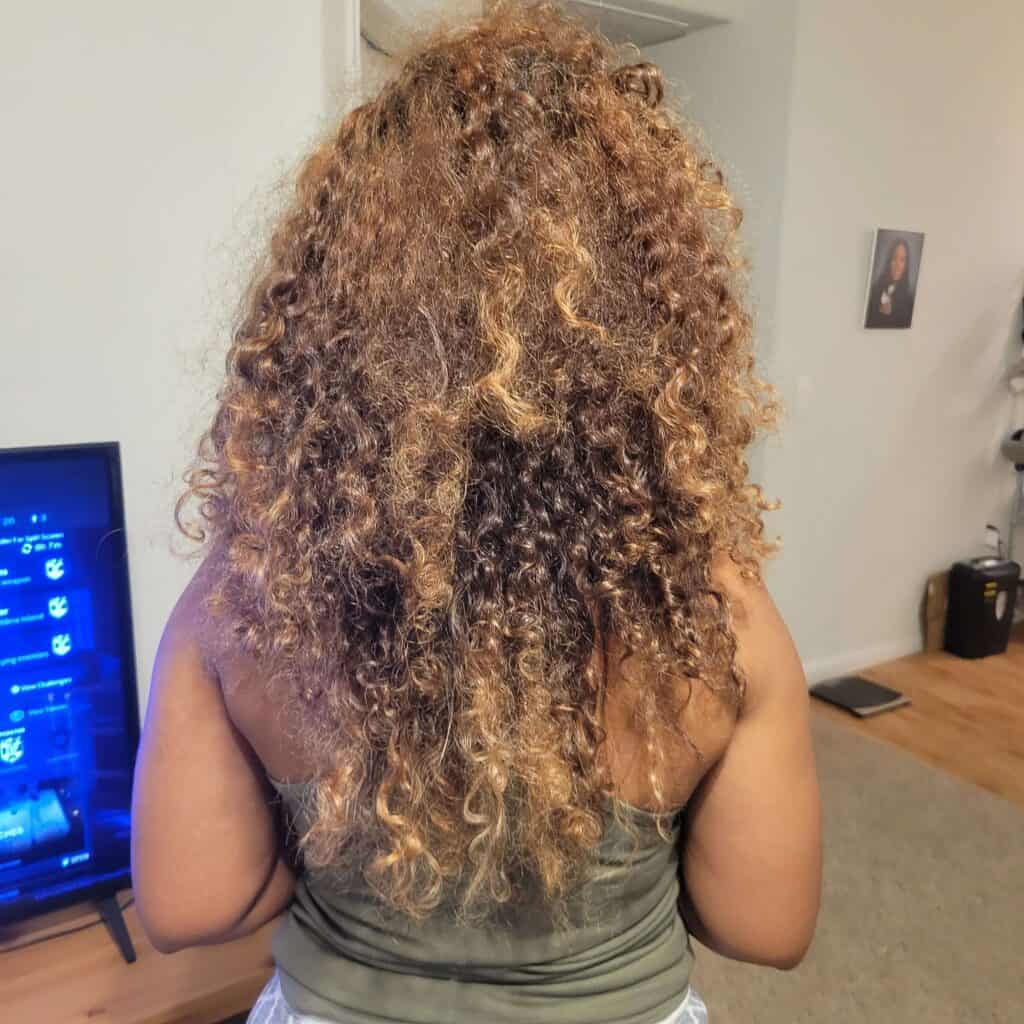 Back view of a woman with highlighted curly hair showing frizz and dryness before using a murumuru hair mask—taken indoors in a casual setting.