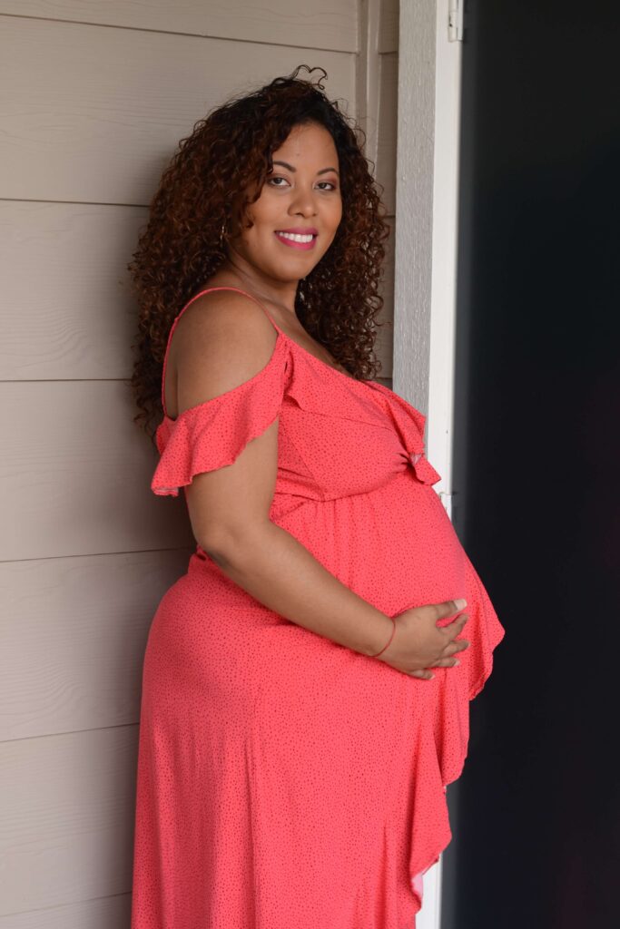 A pregnant woman wearing a flowing pink dress cradling her baby bump while standing in a softly lit room, surrounded by a serene atmosphere