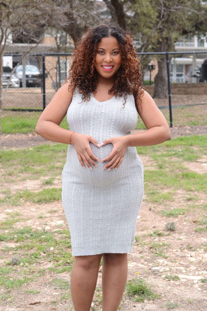 A pregnant woman in a gray dress standing facing forward, forming a heart shape with her hands over her baby bump during a home maternity photoshoot.