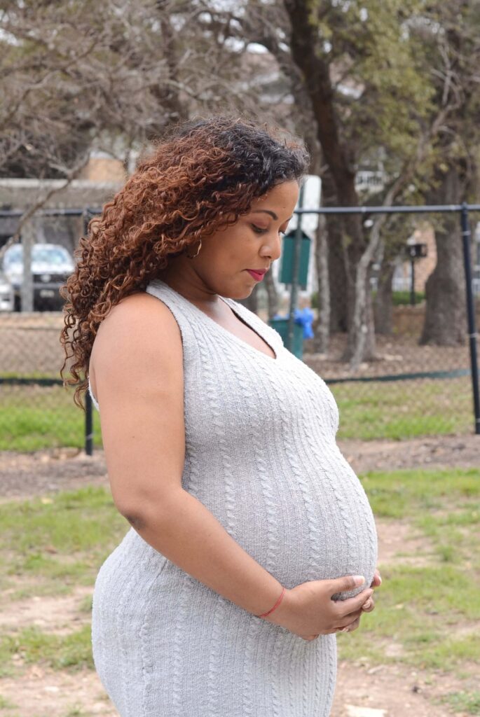 A close-up of a pregnant woman in a gray dress with her hands gently tucked under her baby bump, looking down lovingly at her belly during a home maternity photoshoot.