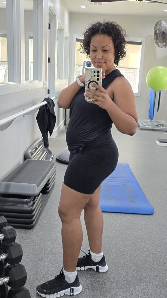 Woman taking a gym mirror selfie in black workout clothes, showing her postpartum body 1.5 years after giving birth, standing confidently with weights and fitness equipment in the background.