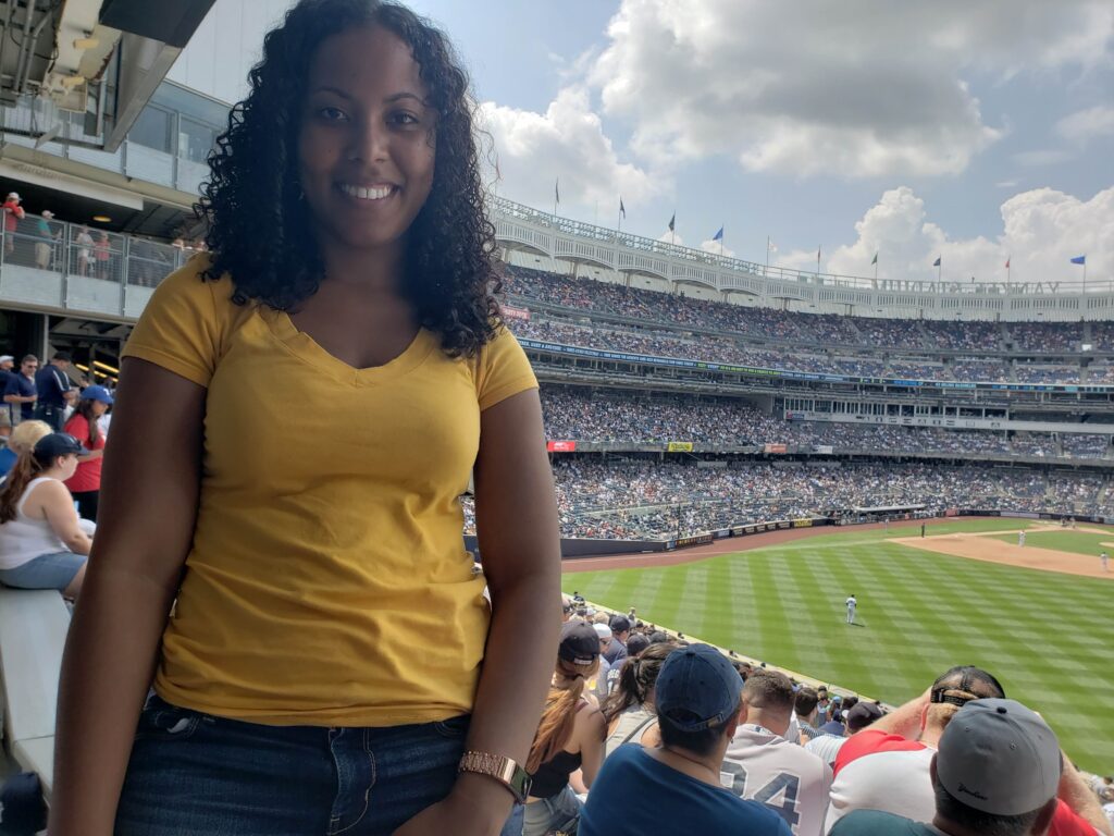 Woman standing in Yankee Stadium, smiling at the camera with the baseball field and stadium seats in the background.