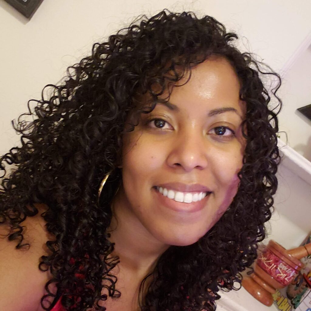 Smiling woman with long, defined black curls standing in a sunlit room, showcasing healthy, voluminous hair.