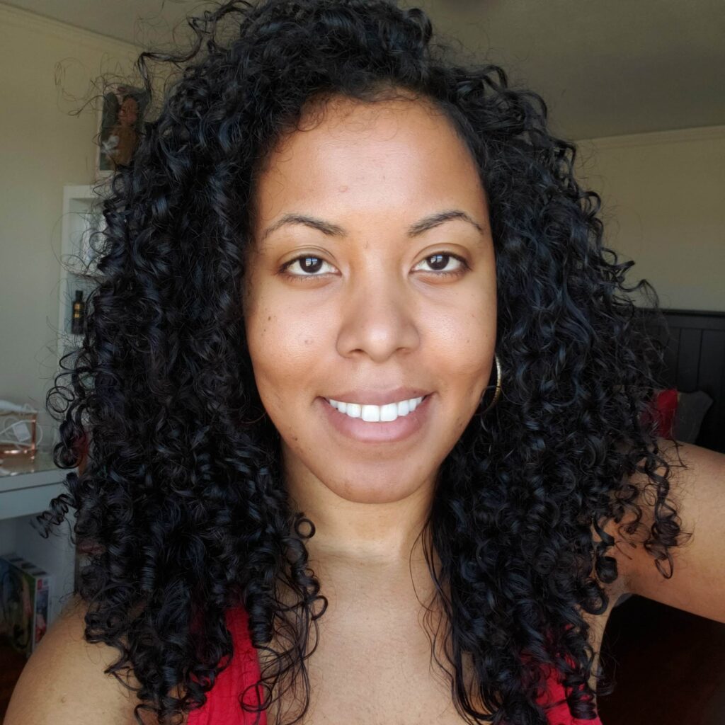 Close-up of a smiling woman with long, shiny, defined curls, taken in warm indoor lighting with kitchen decor in the background.