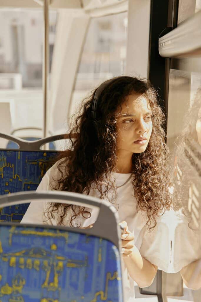 Young woman with loose, 3A curly hair sitting on a bus, wearing headphones and looking out the window. Her curls are elongated, S-shaped, and fall in soft waves — an example of Type 3A curls.