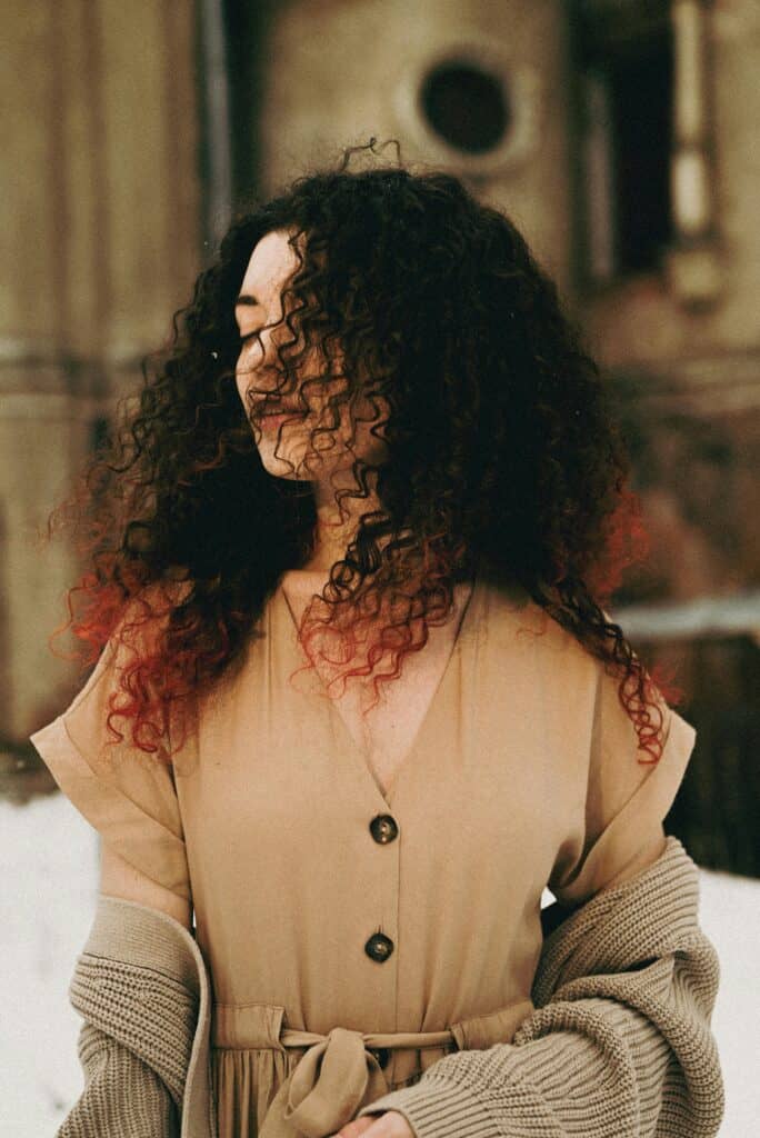 Woman with Type 3B curly hair standing outdoors, her curls forming defined spirals with noticeable volume. Some strands are dyed red at the ends, highlighting the springy 3B curl pattern.