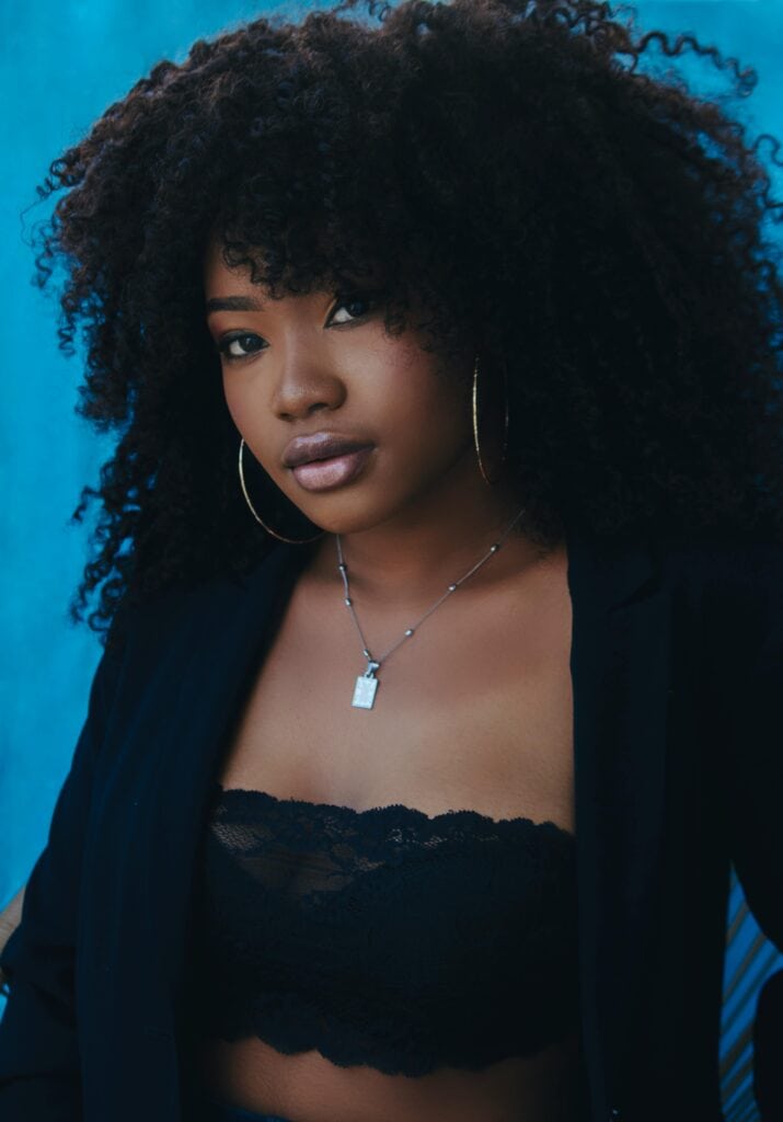 Woman with voluminous 4A natural curls wearing hoop earrings and a black outfit against a blue background, showing defined coily texture and healthy shine.