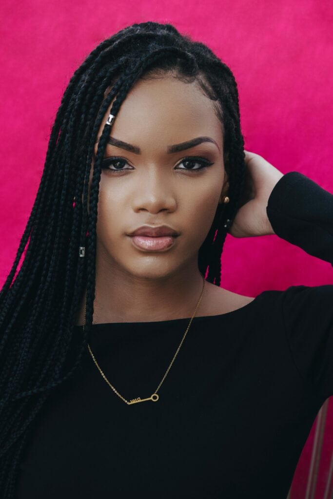 Woman with long box braids wearing a black top and gold necklace against a pink background, holding her hair back confidently.