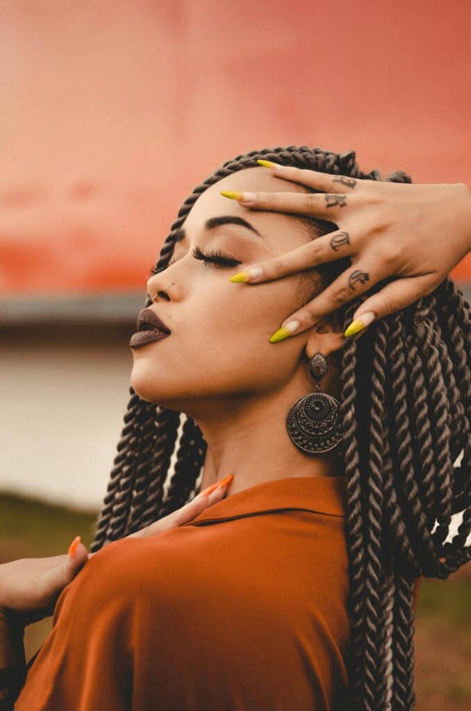 Woman with long two-strand twist braids wearing statement earrings and an orange blouse, posing outdoors with bold makeup and nails.
