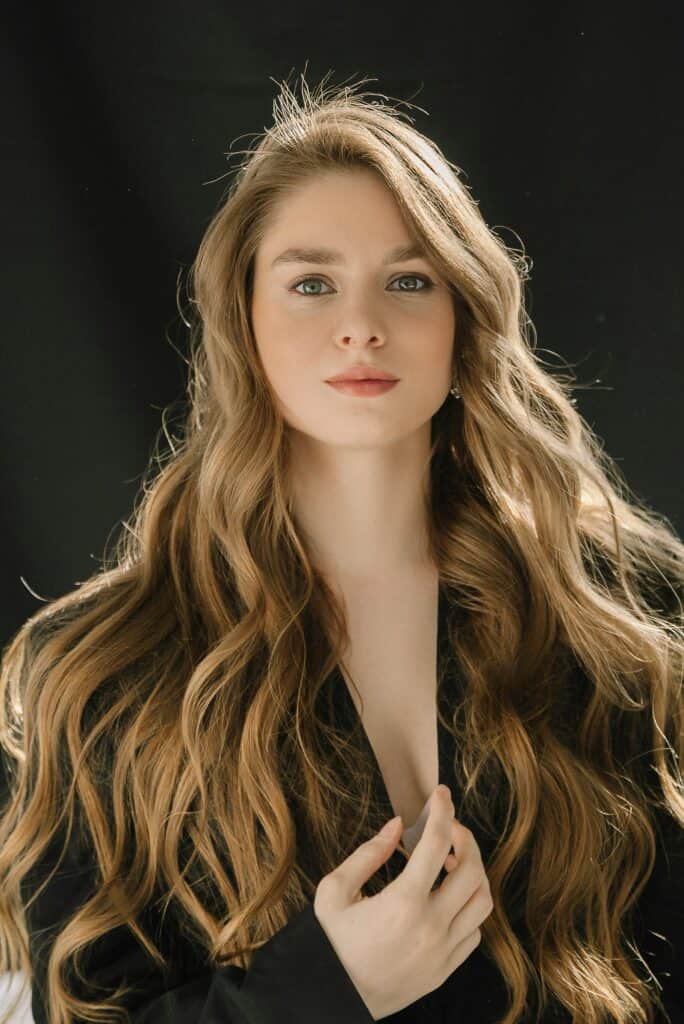Woman with long golden wavy hair wearing a black outfit against a dark background.