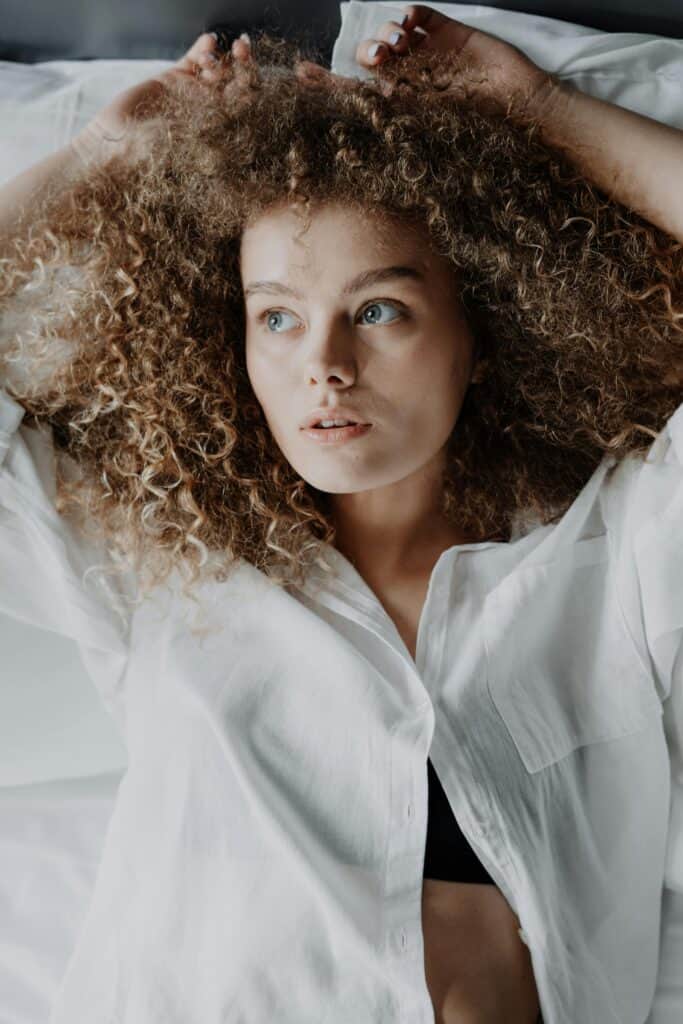 Woman with blonde 3B curls lying on a bed wearing a white shirt, looking to the side.