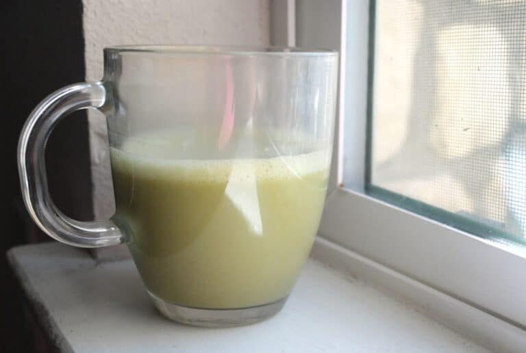 Homemade Jade Leaf matcha latte in a clear glass mug by a window, showing the creamy frothy top and light green color.