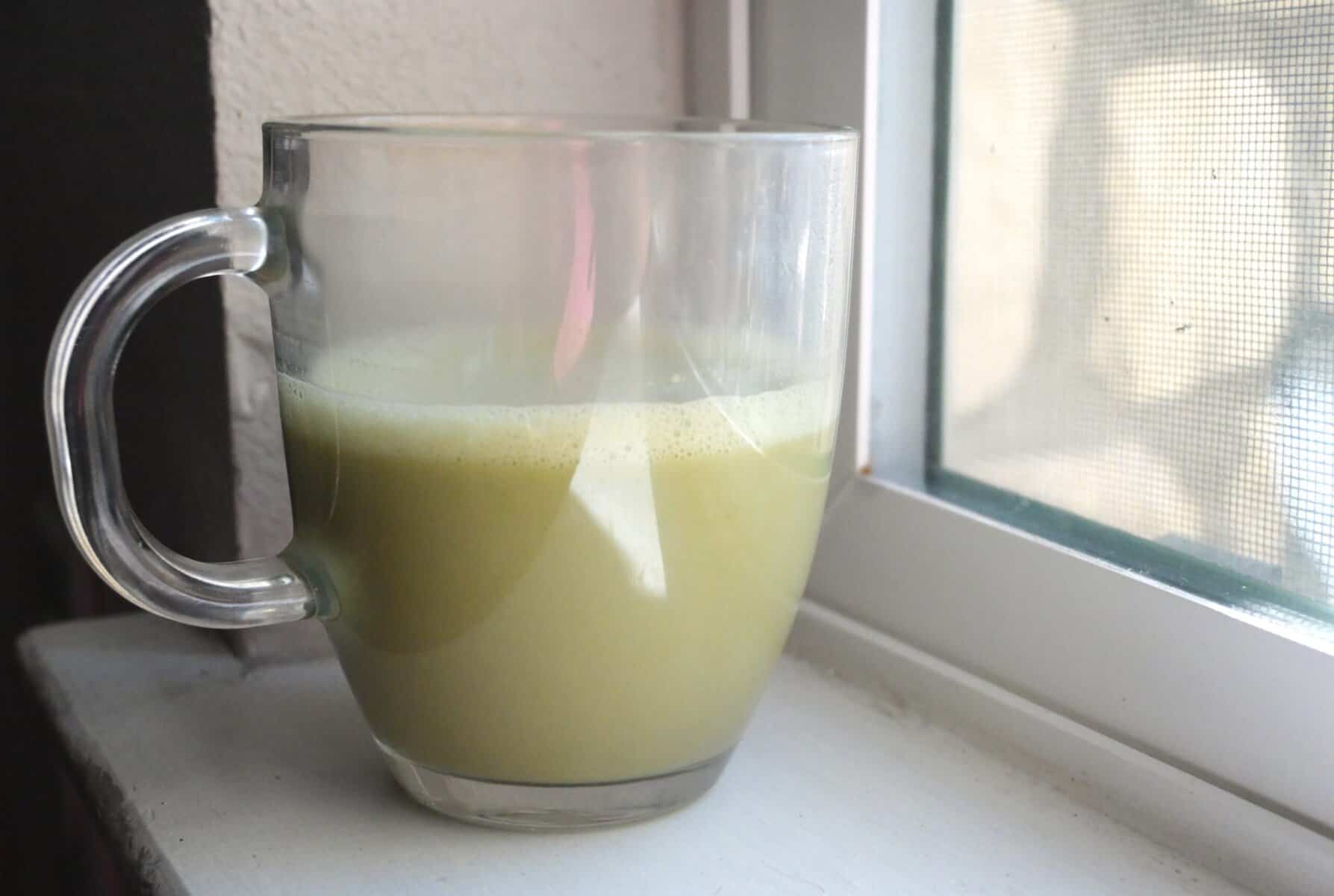 Homemade Jade Leaf matcha latte in a clear glass mug by a window, showing the creamy frothy top and light green color.