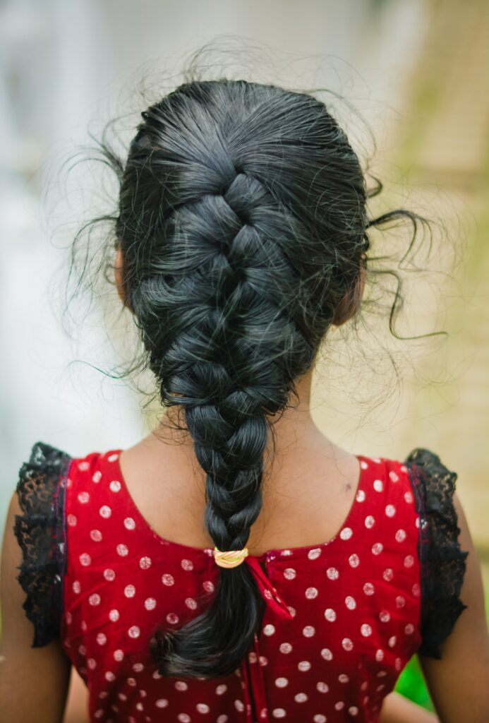 Back view of a school-age girl wearing a long braid protective hairstyle with a few loose strands around the hairline.