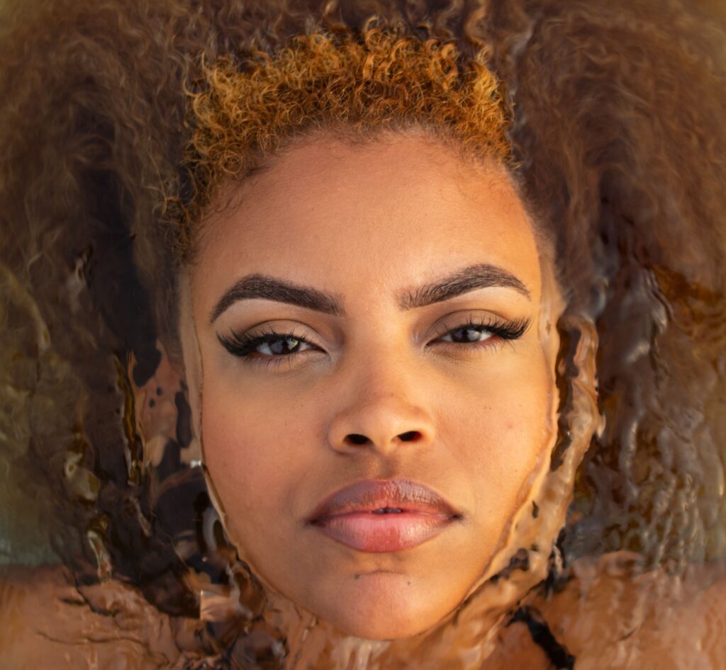Close-up portrait of a woman with curly hair surrounded by water, illustrating how hair porosity affects moisture absorption and retention.