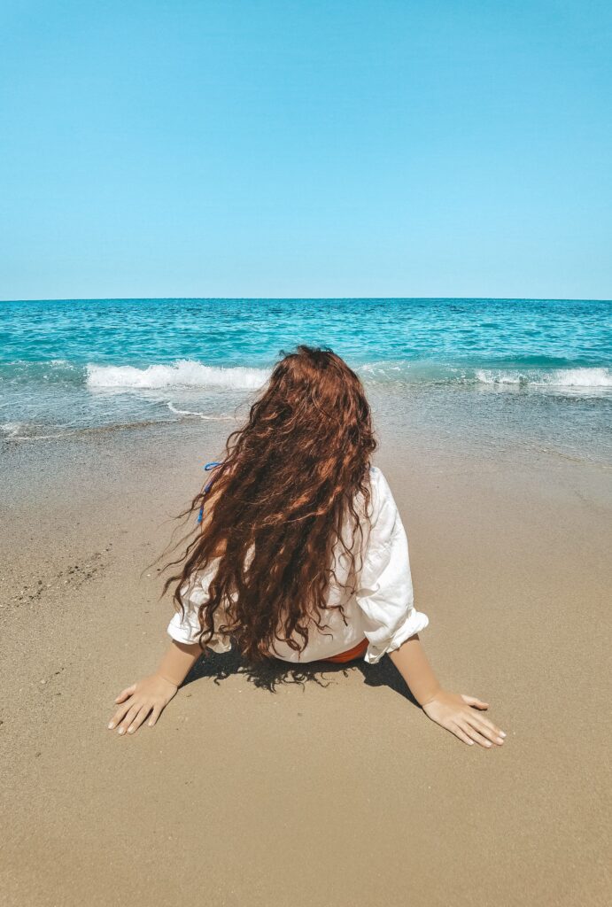 Woman with long wavy hair sitting on the beach facing the ocean.